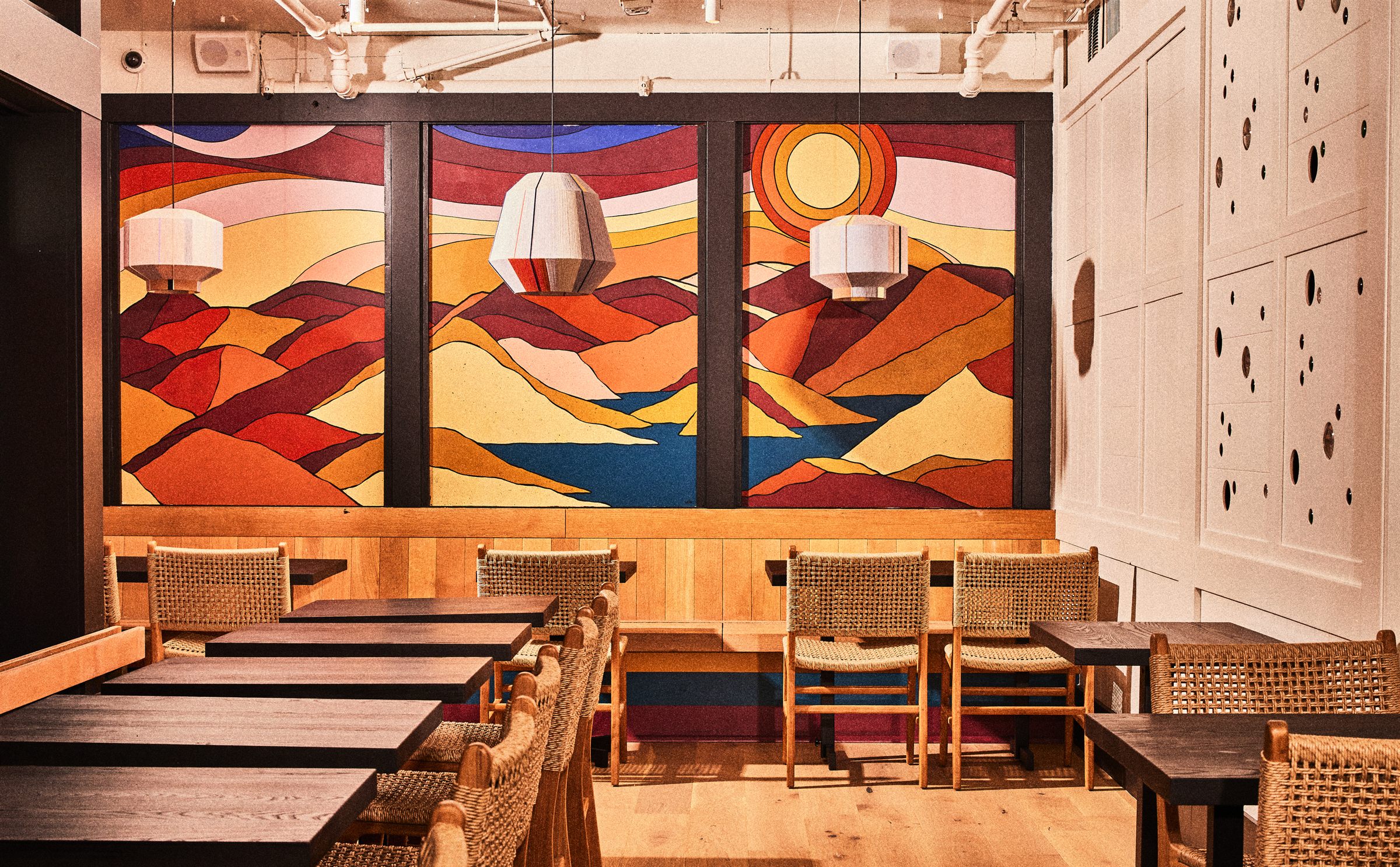 Warm restaurant dining room with mural artwork, pendant lights, and woven chairs